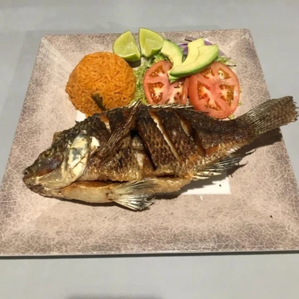 Whole fried fish with Mexican rice, sliced tomatoes, avocado, and lime wedges on a plate.