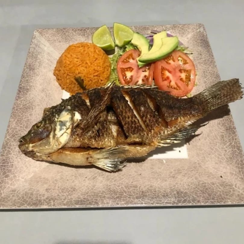 Whole fried fish with Mexican rice, sliced tomatoes, avocado, and lime wedges on a plate.